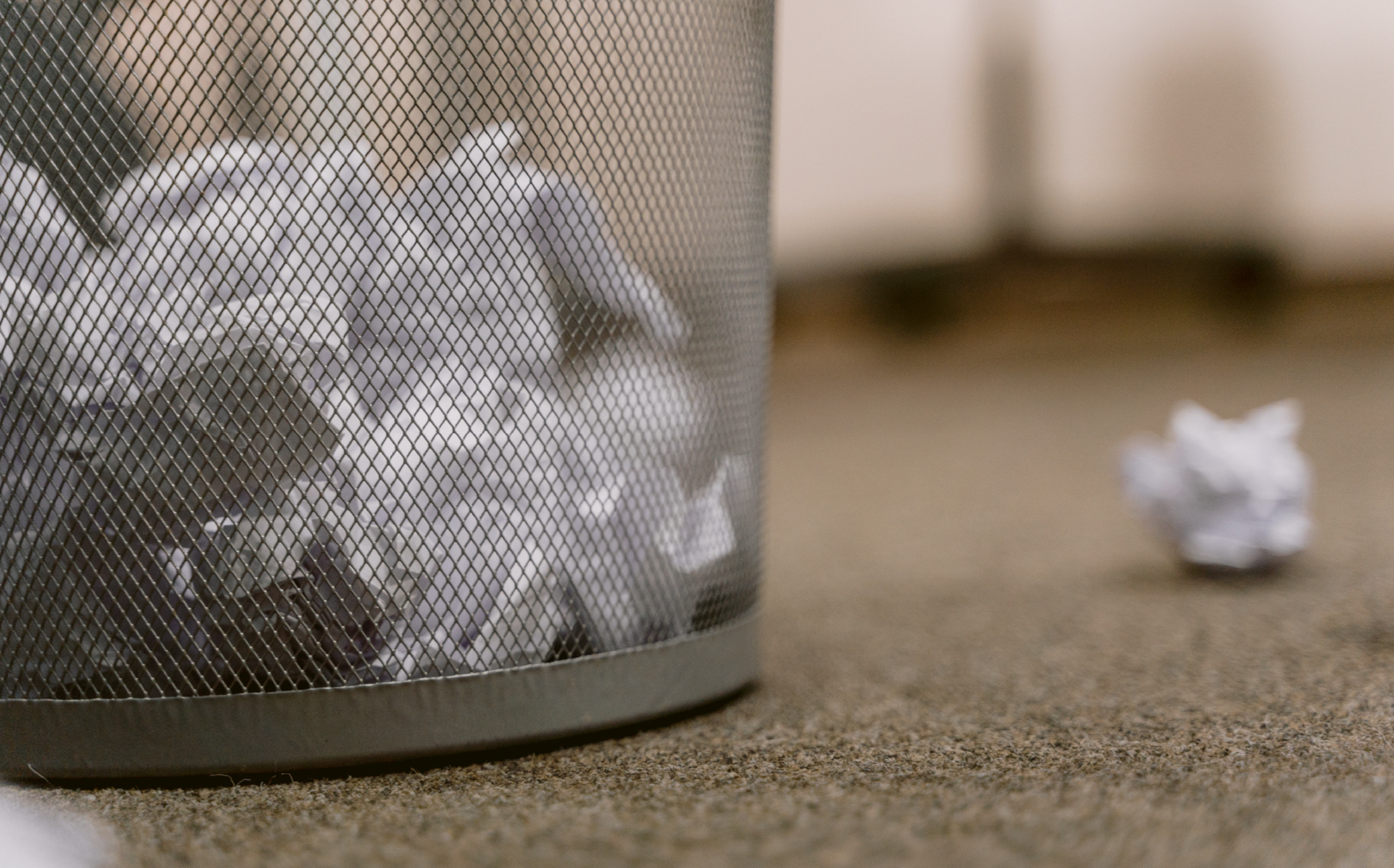 crumbled paper on the floor next to a wire mesh wastebasket with other trash inside of it